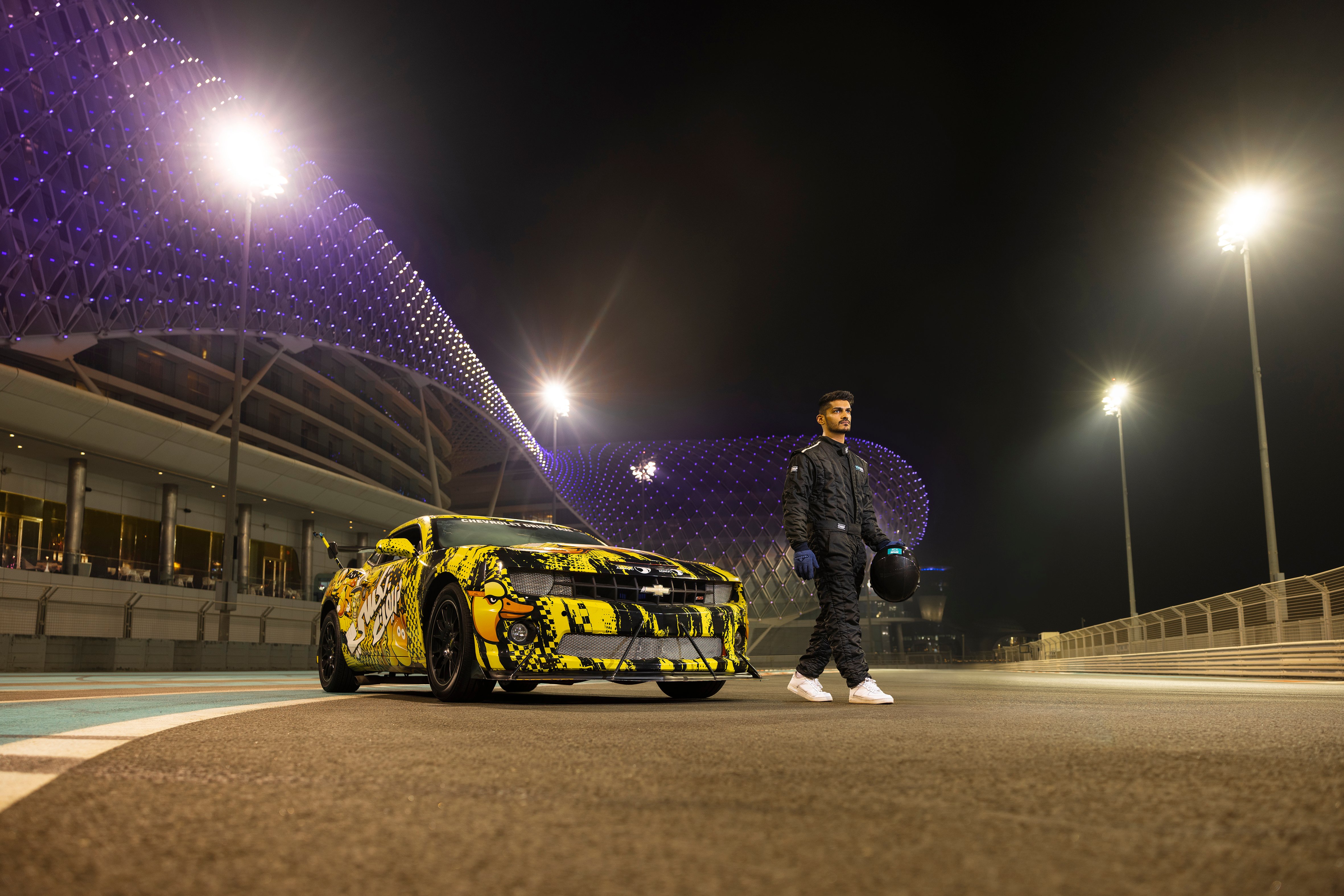 Race car driver walking past custom sports car at Yas Marina Circuit in Abu Dhabi under night lights.