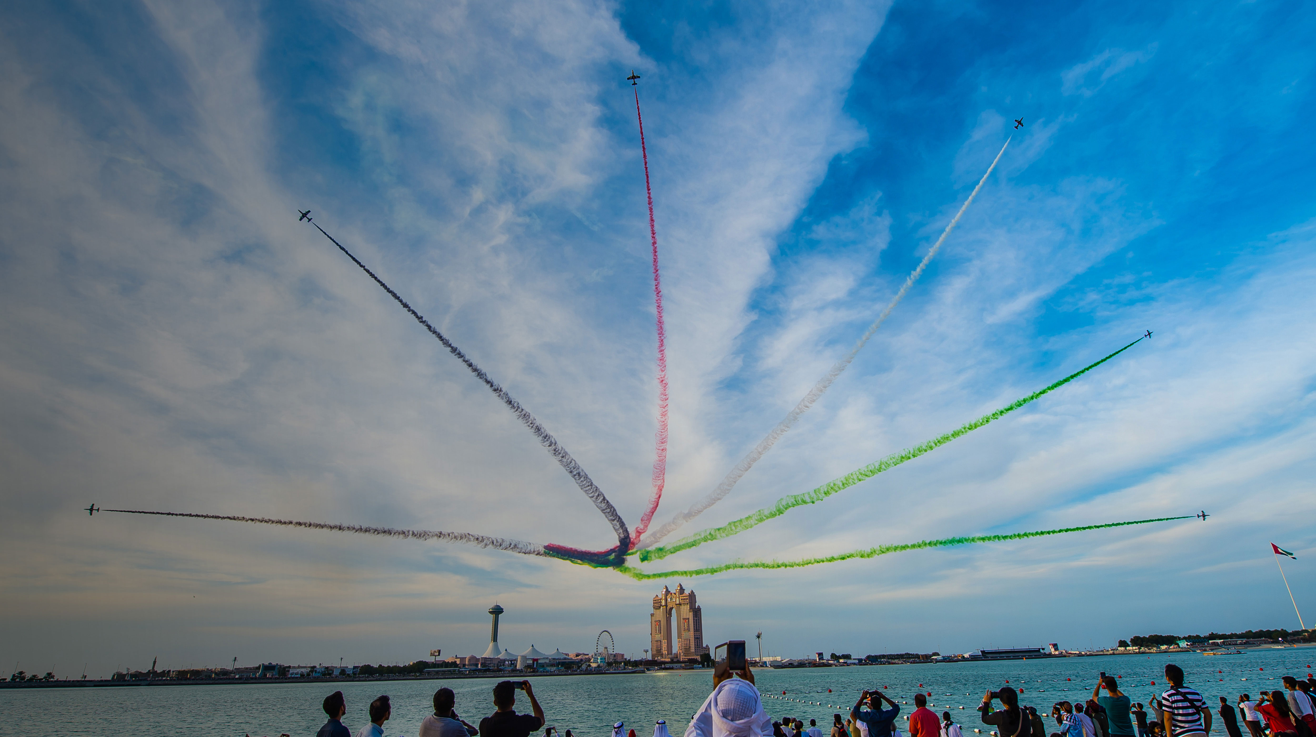 Spectators watch jets perform an airshow over Abu Dhabi skyline with trails of UAE flag colors