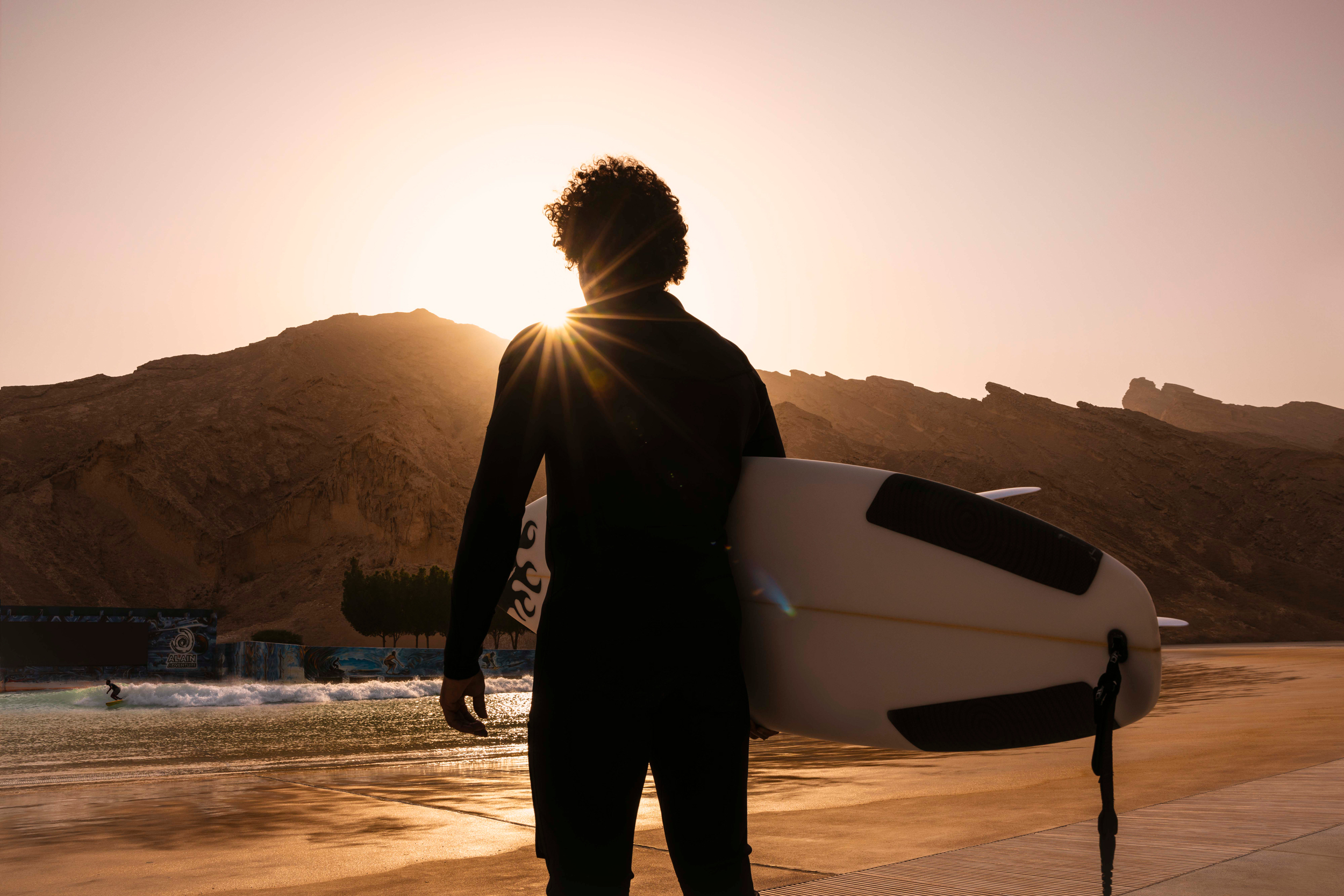 Surfer holding a board at sunrise near man-made waves in Abu Dhabi with rocky mountains in the background.