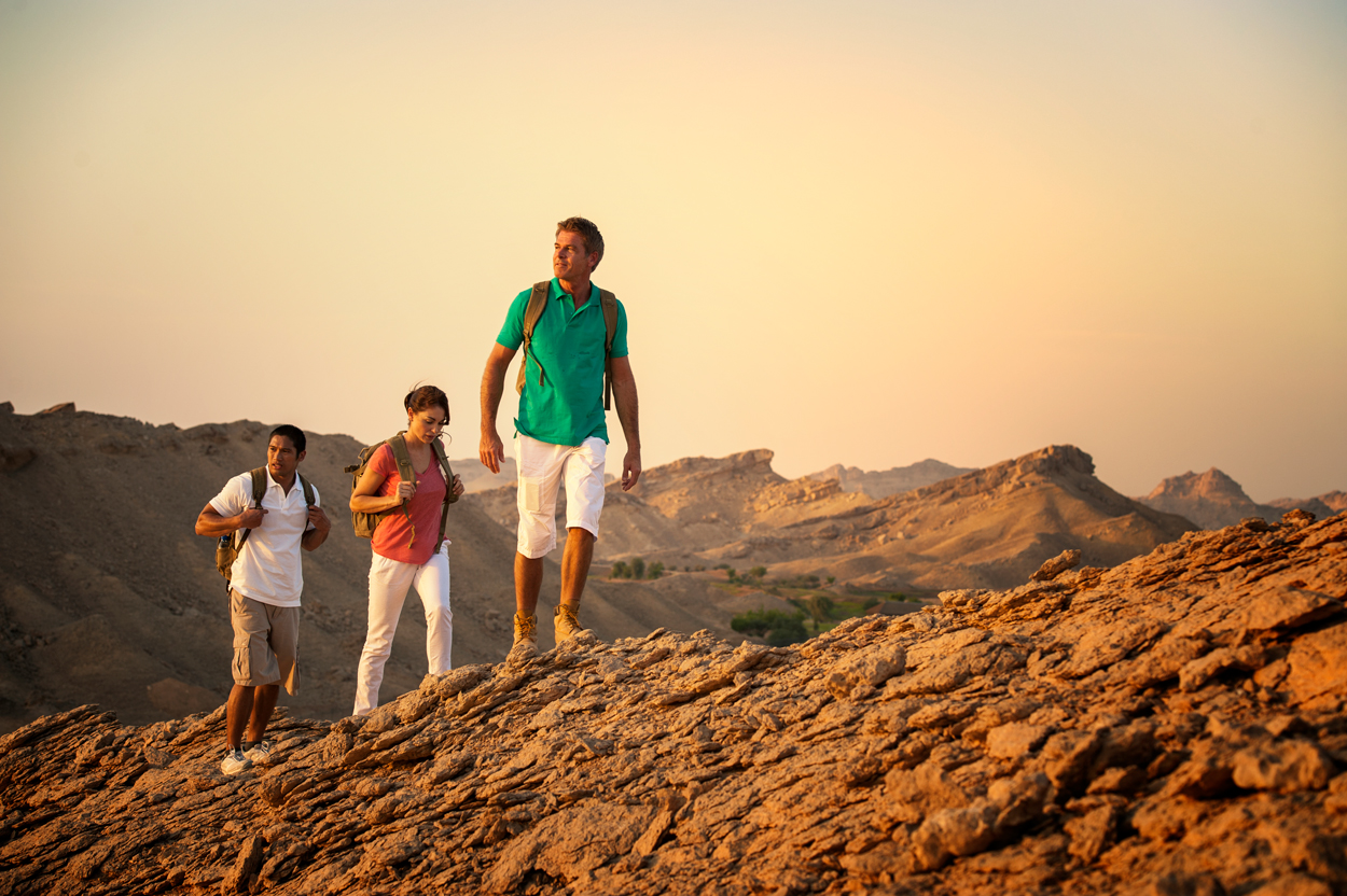 Three hikers walking across rocky terrain at sunset in a desert mountain landscape.