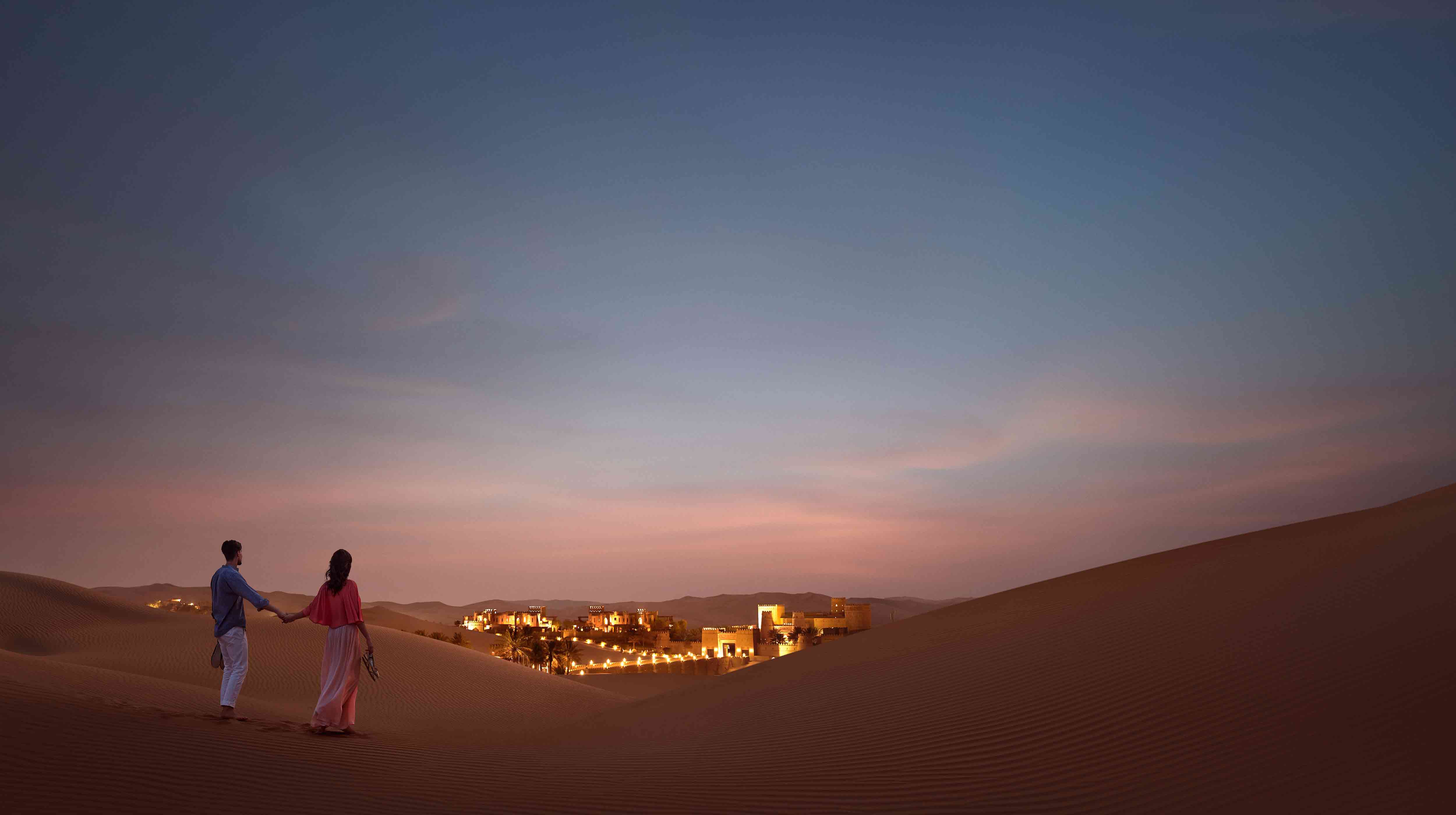 Una coppia ammira al tramonto il complesso architettonico del Qasr Al Sarab Desert Resort by Anantara immerso tra le dune del deserto di Abu Dhabi