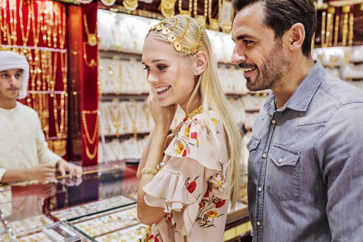 Smiling couple shopping for gold jewelry at a traditional market, with the woman trying on a decorative gold headpiece.